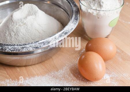 Cooking or baking background with flour in bowl, glass of flour and two eggs on wooden table Stock Photo