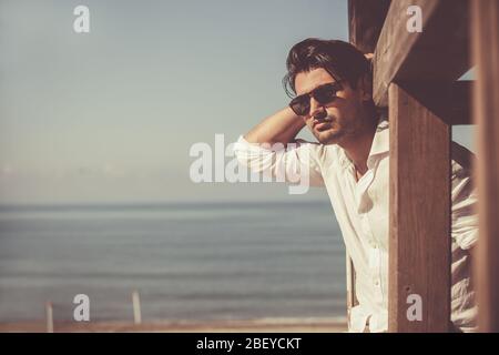 Young attractive man with sunglasses looking out over the sea during the summer. He looking forward, dressed in a white shirt and leaning on a wooden Stock Photo