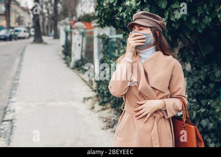 Woman coughing wears reusable mask outdoors during coronavirus covid-19 pandemic. Girl feeling sick. Keep distance Stock Photo