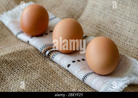 Three brown chicken eggs on burlap. A line of raw farm eggs on a rushnyk. A rolled towel with eggs on burlap. Angled side view. Close-up. Selective fo Stock Photo