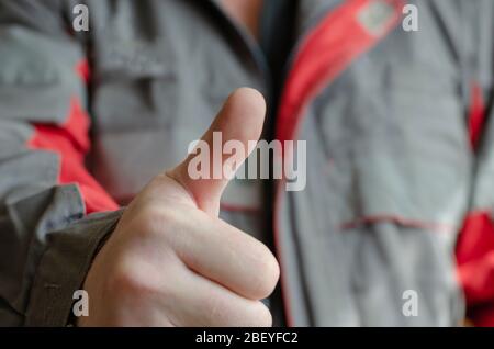 A man shows the camera a thumb up gesture. Mechanic in a red-gray working uniform with a raised thumb on his hand. Wearing special clothing in the wor Stock Photo