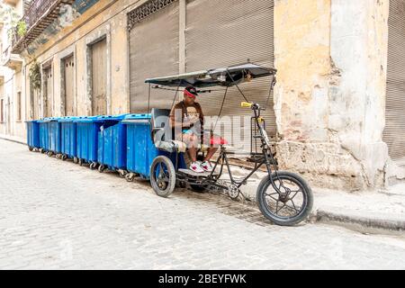 Uncluttered view of a Cuban rickshaw taxi with male taxi driver counting his money with a train of bins behind him, Havana old town, Cuba Stock Photo