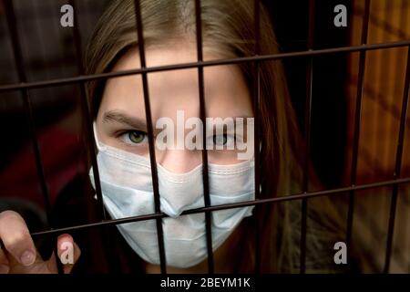angry young woman in mask looks frowning through insulation grid. Concern for future with the spread of deadly virus. Stock Photo