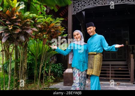 A portrait of young couple of malay muslim in traditional costume during Aidilfitri celebration showing welcome greeting gesture by traditional wooden Stock Photo