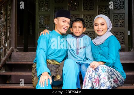 A portrait of young couple of malay muslim in traditional costume with his son during Eid al-Fitr celebration by traditional wooden house. Raya and Mu Stock Photo