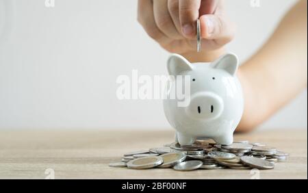 Money savings concepts hand holding coin to put in piggy bank to spend on expenses such as savings, tourism, investment, emergency, retirement on wood Stock Photo