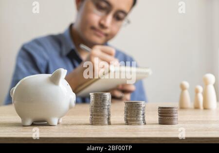 Money savings concepts  piggy bank and stack coins with blur man who's taking notes about saving money for family and family wooden dolls.on wooden ta Stock Photo