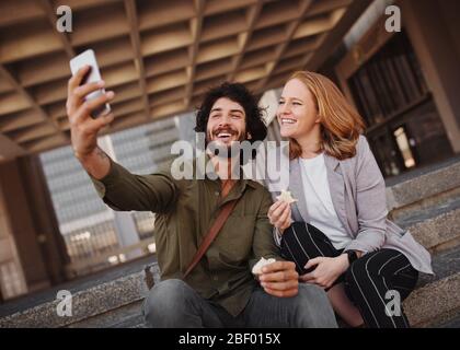 Happy professional couple taking selfie using smartphone while eating sandwich for lunch during break Stock Photo
