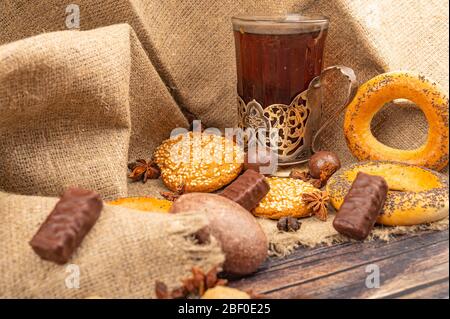 A faceted glass of tea in a vintage Cup holder, bagels, cookies and chocolate gingerbread on a background of rough homespun fabric. Close up Stock Photo