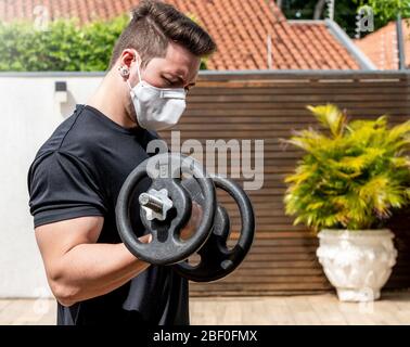 Portrait of a man wearing face mask doing exercises at home during quarantine. Man doing biceps curl with a dumbbell alone at home. Stock Photo