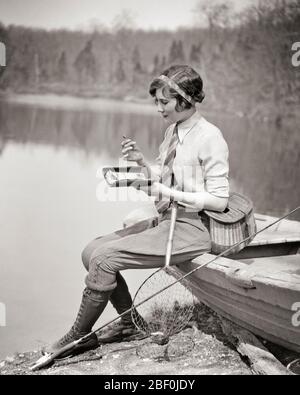 1920s YOUNG ATTRACTIVE SPORTS WOMAN WELL EQUIPPED WITH ROD REEL CREEL SITTING ON BOAT BY LAKE SELECTING FISHING FLY FROM WALLET - a1458 HAR001 HARS RURAL HEALTHINESS COPY SPACE FULL-LENGTH ATHLETIC HOOK ROD CONFIDENCE B&W CATCHING SKILL ACTIVITY AMUSEMENT HAPPINESS CHEERFUL HOBBY LEISURE STRENGTH INTEREST CHOICE HOBBIES KNOWLEDGE RECREATION PASTIME REEL PLEASURE BY ON NECKTIE SMILES JOYFUL STYLISH ATTRACTIVE JODHPURS RELAXATION YOUNG ADULT WOMAN AMATEUR BLACK AND WHITE CAUCASIAN ETHNICITY CREEL ENJOYMENT HAR001 OLD FASHIONED SELECTING Stock Photo