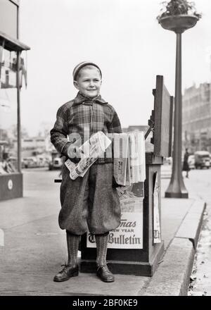 1930s SMILING BOY SELLING NEWSPAPERS FROM CORNER STAND IN THE CITY WEARING WOOLEN KNICKERS JACKET GLOVES KNIT CAP - b3768 HAR001 HARS LIFESTYLE SATISFACTION UNITED STATES COPY SPACE FULL-LENGTH BULLETIN UNITED STATES OF AMERICA MALES CORNER NEWSPAPERS CONFIDENCE B&W KNIT NORTH AMERICA WINTERTIME GOALS NORTH AMERICAN SELLING HAPPINESS CUSTOMER SERVICE PA OCCUPATIONS NEWSBOY WOOLEN GROWTH JUVENILES PRE-TEEN PRE-TEEN BOY BLACK AND WHITE CAUCASIAN ETHNICITY HAR001 OLD FASHIONED Stock Photo