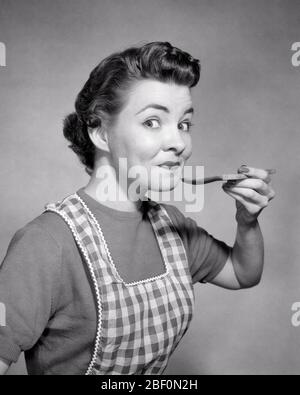 1950s SMILING WOMAN HOUSEWIFE WEARING CHECKERED APRON LOOKING AT CAMERA WITH RAISED EYEBROWS TASTING FROM WOODEN KITCHEN SPOON - g3729 CLE003 HARS COPY SPACE FRIENDSHIP HALF-LENGTH LADIES PERSONS CARING EXPRESSIONS B&W EYE CONTACT HOMEMAKER HAPPINESS HOMEMAKERS CHEERFUL EXCITEMENT PRIDE TASTE TASTING EYEBROWS HOUSEWIVES SMILES APPROVING JOYFUL STYLISH CHECKERED MID-ADULT MID-ADULT MAN SAMPLE BLACK AND WHITE CAUCASIAN ETHNICITY FLAVOR OLD FASHIONED Stock Photo