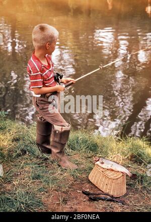 1960s BLOND BOY WEARING RED T-SHIRT RUBBER BOOTS STANDING FISHING ON STREAM  BANK WITH ROD AND REEL CREEL AND THREE CAUGHT FISH - ka707 LAN001 HARS JOY  LIFESTYLE SATISFACTION STRIPED RURAL COPY