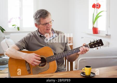 Senior man playing guitar at home in a bright living room with a cup of espresso on the table Stock Photo