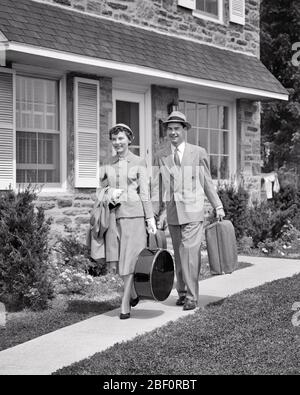 1950s SMILING COUPLE WALKING TOGETHER CARRYING LUGGAGE LEAVING SUBURBAN HOUSE GOING ON VACATION TRIP - s1750 HAR001 HARS VACATION STRONG PLEASED JOY LIFESTYLE SATISFACTION CELEBRATION FEMALES MARRIED SPOUSE HUSBANDS HEALTHINESS HOME LIFE FULL-LENGTH LADIES PERSONS INSPIRATION CARING MALES B&W EYE CONTACT TIME OFF SUIT AND TIE DREAMS HAPPINESS CHEERFUL ADVENTURE TRIP GETAWAY EXTERIOR RECREATION ON HOLIDAYS SMILES CONCEPTUAL ESCAPE JOYFUL STYLISH PERSONAL ATTACHMENT AFFECTION EMOTION MID-ADULT MID-ADULT MAN MID-ADULT WOMAN RELAXATION TOGETHERNESS VACATIONS WIVES BLACK AND WHITE Stock Photo