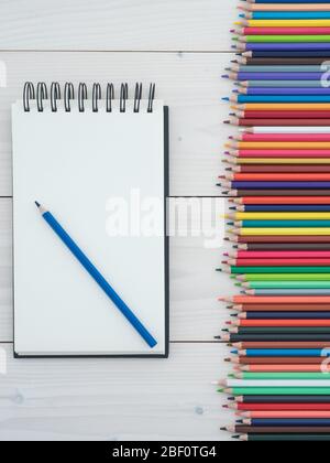 Set of colored pencils arranged in a row and notebook on wooden background. top view Stock Photo