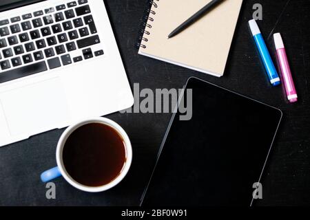 View from above of modern workplace with cup of tea, laptop, tablet, pens and notebook on black wooden table Stock Photo