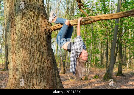 Young woman hangs upside down with crossed legs from a thick branch. Side view. Location: Germany, North Rhine Westphalia, Hoxfeld Stock Photo