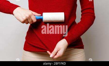 Woman hand using a sticky roller to clean fabrics - red woolen turtleneck from dust, hair, lint and fluff, front view, close up. Grey background. Stock Photo