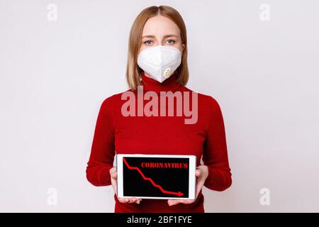 European woman wearing a face mask, showing red arrow moving down, recession process in the economy on digital tablet, isolated on grey background. Co Stock Photo