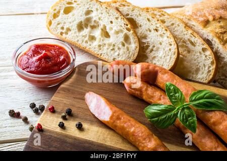 Thin smoked sausages near ketchup and sliced bread on a brown wooden cutting board. Traditional sausages in daily nutrition. Front view. Stock Photo