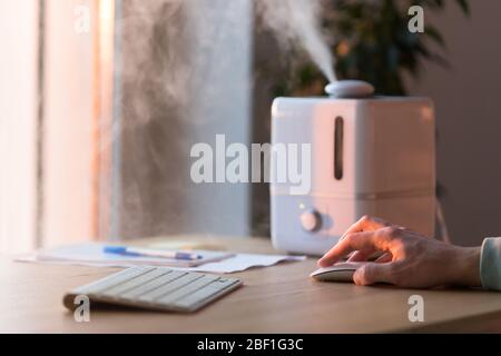 Man working on computer, using pc mouse near the aroma oil diffuser on the table, steam from humidifier, selective focus. Humidification of air in the Stock Photo