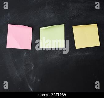 three blank paper multi-colored square stickers are stuck on a black chalk board Stock Photo