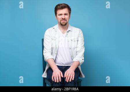 Thoughtful stressed young man on a chair sitting alone on blue wall. Stock Photo