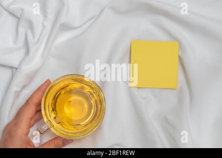 Woman hand holding cup of tea and empty sticky note on white bed background, mockup copy space Stock Photo