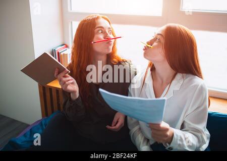 Two young women are tired and having fun. They imitate a mustache with the help of multi-colored pencils. Two red-haired students study at home or in Stock Photo
