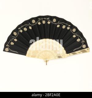 Pleated fan. Pleated fan with a black silk leaf with a simple meandering floral vine embroidered with white silk and appliquéd with white machine made lace flowers. Drilled and pierced bone sticks. Mother-of-pearl washer and metal loop at the rivet. Stock Photo