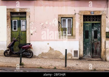 Maroon scooter parked in front of rustic pink building with two green doors in Lisbon's Alfama district Stock Photo
