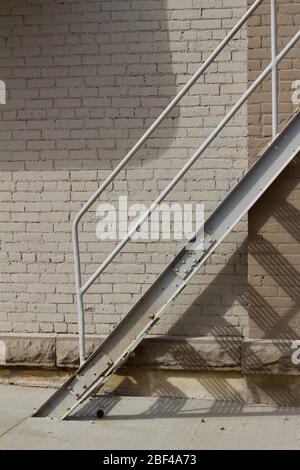 Close up view of an industrial looking metal fire escape stairs attached to a vintage gray brick wall building surface, casting shadows on a sunny day Stock Photo