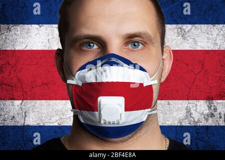 Young man with sore eyes in a medical mask painted in the colors of the national flag of Costa Rica. Coronovirus disease  COVID-19 concept.  Man is af Stock Photo