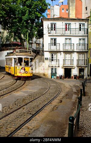 Yellow electric tram 28 descends a hill past a white house with balconies in the Alfama district of Lisbon Stock Photo
