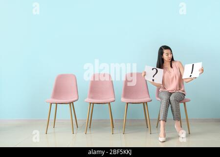 Young woman holding paper sheets with question and exclamatory marks while sitting on chair indoors. Job interview concept Stock Photo