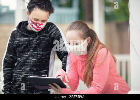 A young man and woman watching a tablet wearing cloth face masks. Covering face in public is recommended by CDC in many countries during to Covid-19 Stock Photo