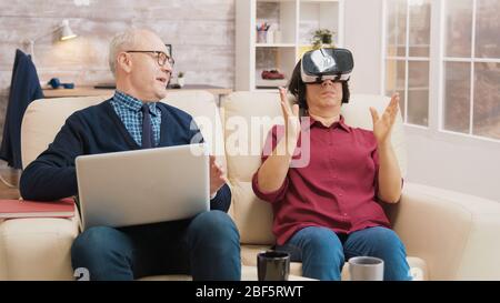 Amazed senior woman while using virtual reality goggles on sofa with her husband next to her using laptop. Stock Photo