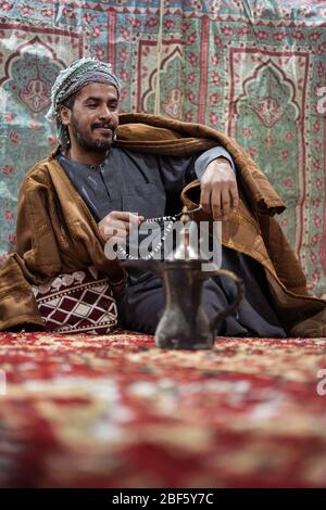 Bedouin man wearing traditional clothes praying with a tasbih while drinking tea on a carpet in the Saudi desert during the night, Saudi Arabia Stock Photo