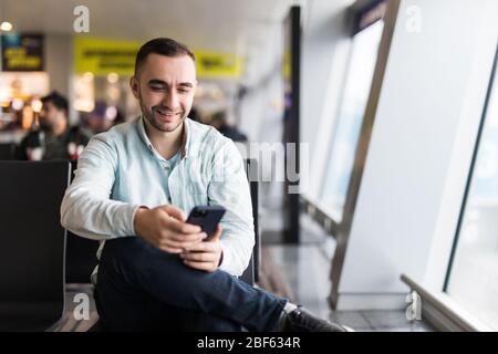 Texting he is fine. Portrait of handsome man in casual wear holding his luggage and messaging through his mobile phone while sitting in the hall of th Stock Photo