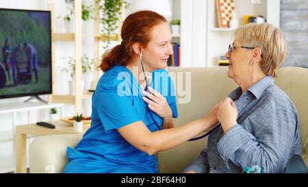 Caucasian female doctor in nursing home listening old woman heartbeat with stethoscope Stock Photo