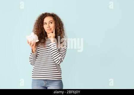 Thoughtful African-American woman with piggy bank on color background Stock Photo