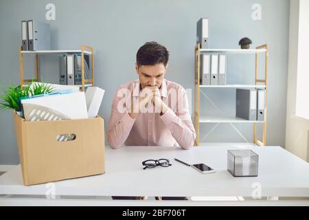 Unemployment. Dismissal. Dismissed businessman is upset with a cardboard box holding his head in his hands while sitting in a company office. Stock Photo