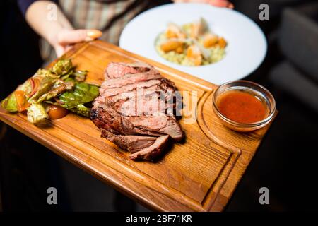 Freshly prepared meat steak sliced into pieces on a wooden board. Served with sauce and green salad. Stock Photo