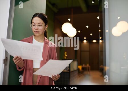 Young Asian businesswoman in eyeglasses examining future project on papers while standing at office Stock Photo