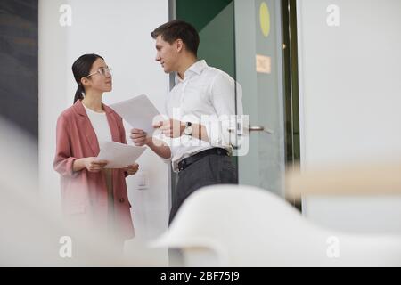 Two colleagues holding documents and talking to each other they discussing business documents while standing in the corridor at office Stock Photo