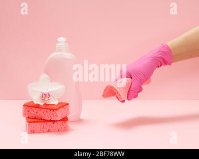 Plastic bottle of dishwashing liquid, red sponges, a white orchid flower and a hand in a rubber glove holding a rag on the pink background. Washing an Stock Photo