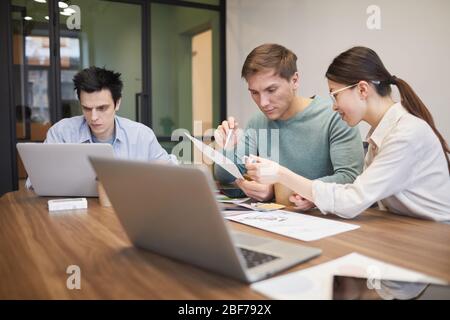 Businessman and businesswoman sitting at the table with laptop and discussing document with their colleague sitting near by and using his laptop at of Stock Photo