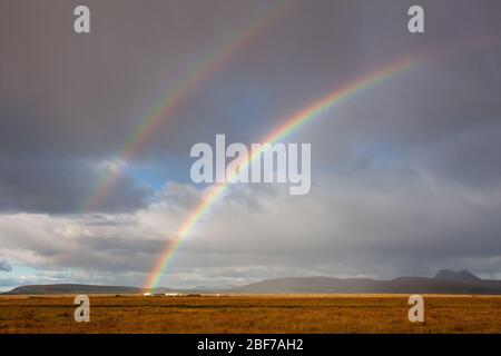 A double rainbow appears over sun-lit yellow fields and in front of a cloudy sky in southern Iceland Stock Photo